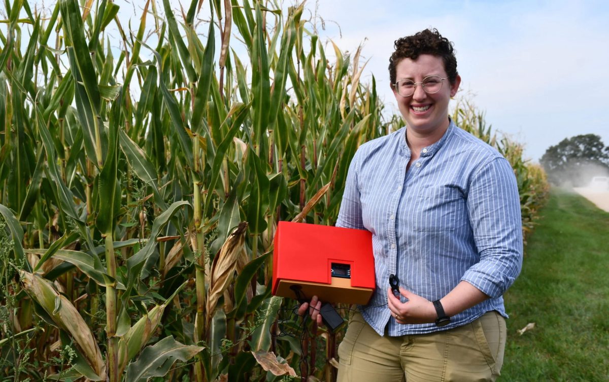 Woman with short hair and glasses holding a red piece of machinery standing in front of corn stalks.