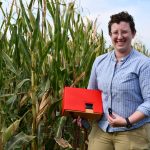 Woman with short hair and glasses holding a red piece of machinery standing in front of corn stalks.