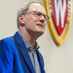 A closeup of a white man with short gray hair and glasses speaking to an audience