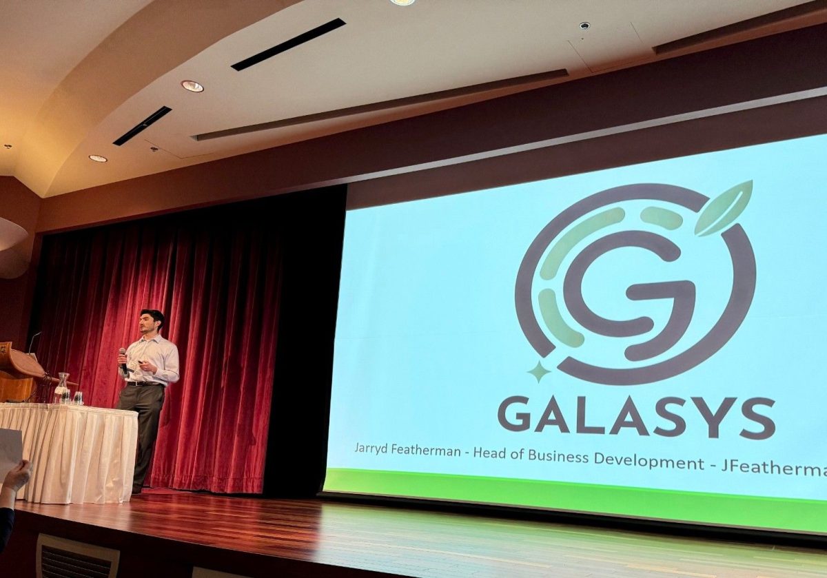 a man stands on a stage with a microphone. the screen behind him reads "Galasys, Jarryd Featherman, head of business development"