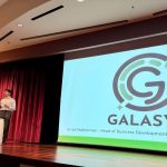 a man stands on a stage with a microphone. the screen behind him reads "Galasys, Jarryd Featherman, head of business development"